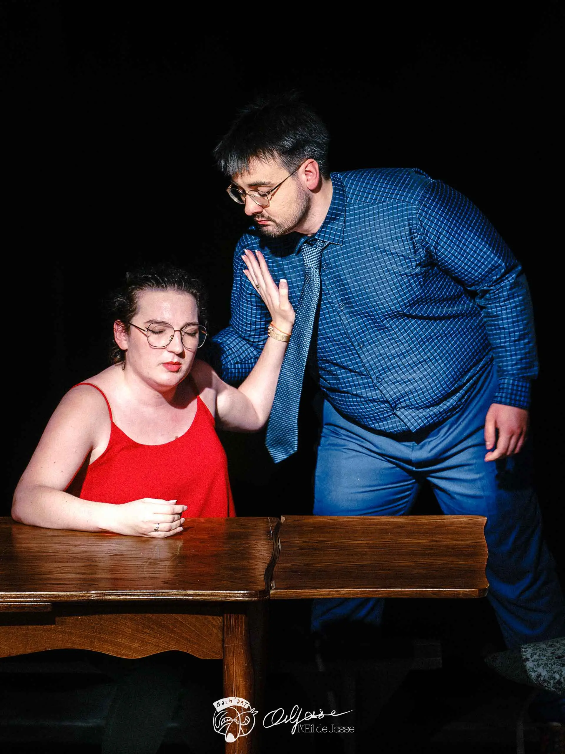 Un comédien en chemise bleue se penche vers une comédienne en débardeur rouge, assise à une table.