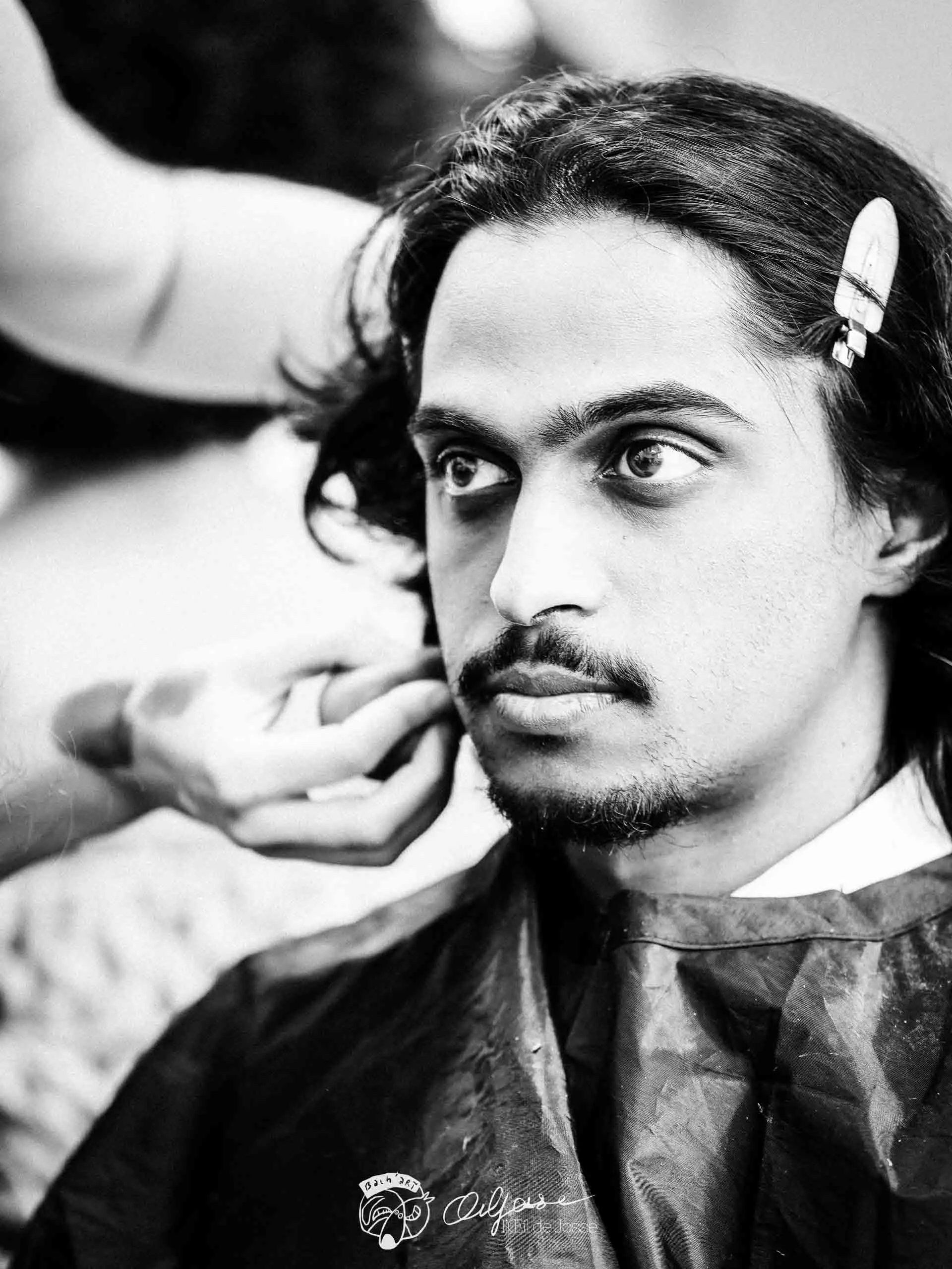 Un comédien aux cheveux longs se laisse maquiller, une barrette dans les cheveux.